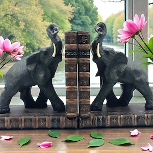May include: A pair of dark gray elephant bookends with raised trunks, flanking two vintage-style books. The bookends sit on a decorative base with the word "VOYAGE" and are surrounded by pink flowers.