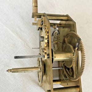 Puede incluir: Un mecanismo de relojer&iacute;a de lat&oacute;n con varios engranajes y una llave de cuerda. El mecanismo est&aacute; montado en una placa de lat&oacute;n de forma rectangular.