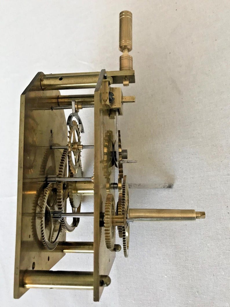 Puede incluir: Un mecanismo de reloj de lat&oacute;n con varios engranajes y una llave de cuerda. El mecanismo est&aacute; montado en una placa de lat&oacute;n y tiene una varilla de lat&oacute;n que se extiende desde el lateral.