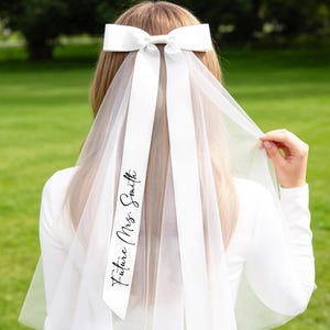 May include: A white bridal veil with a large bow at the top. The veil features long ribbons with the text "Future Mrs. Smith" printed in black script. The bride is wearing a white dress.
