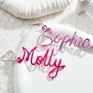 Puede incluir: Dos llaveros con nombres de alambre rosa, uno que deletrea "Sophia" y el otro "Molly". Los llaveros están sobre una superficie blanca con un objeto decorativo blanco en el fondo. Los llaveros tienen cierres plateados.