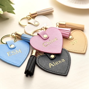 Può includere: Una collezione di portachiavi a forma di cuore in vari colori, tra cui rosa, blu, nero e beige. Ogni portachiavi presenta un anello e un nappa in metallo dorato. Alcuni portachiavi sono personalizzati con nomi o iniziali in lettere dorate.