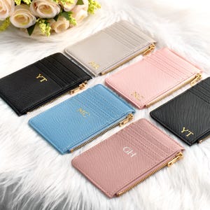 Könnte beinhalten: Eine Auswahl an personalisierten Kartenhaltern in verschiedenen Farben, darunter Schwarz, Hellgrau, Hellblau und Rosa. Jeder Kartenhalter hat einen goldenen Reißverschluss und goldene Initialen. Die Kartenhalter liegen auf einer weißen, flauschigen Oberfläche.