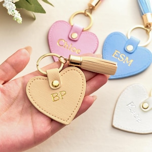 Könnte beinhalten: Herzförmige Schlüsselanhänger in Pastellfarben: Rosa, Beige und Blau. Jeder Schlüsselanhänger hat goldfarbene Hardware und einen goldenen Initialen- oder Namensaufdruck. Der beige Schlüsselanhänger wird in einer Hand gehalten. Weitere Schlüsselanhänger sind in Hellblau, Rosa und Weiß.
