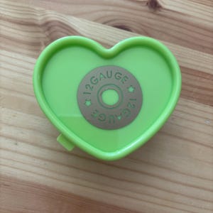 May include: A heart-shaped, lime green container with a circular, tan-coloured design in the centre. The design features the text "12 GAUGE" in a circular pattern. The container is closed and sits on a wooden surface.