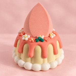 May include: A peach-colored, teardrop-shaped makeup sponge resting on a pale yellow, fluted base. The base is decorated with pink icing drips, small white and pink flowers, green leaves, and a border of white rounded icing beads at the bottom.