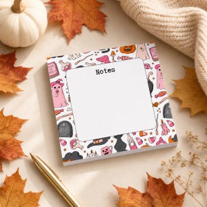 May include: A square notepad with a white center and the word Notes printed at the top. The border features a repeating pattern of pink ghosts, black cats, orange pumpkins, tombstones, mushrooms, candy, and spell books on a white background.