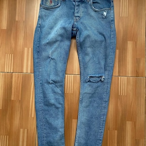 Puede incluir: Vaqueros de mezclilla azul con detalles desgastados, incluyendo una rodilla rasgada y áreas descoloridas. Cuenta con un pequeño parche con el logo en el bolsillo delantero y cierre de botón. Los vaqueros tienen un diseño clásico de cinco bolsillos.