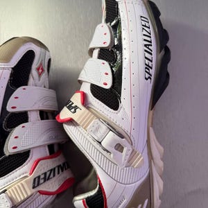 May include: White and black Specialized cycling shoes with three adjustable straps. The shoes have a red and beige accent colour scheme, with the word "SPECIALIZED" printed on the side.