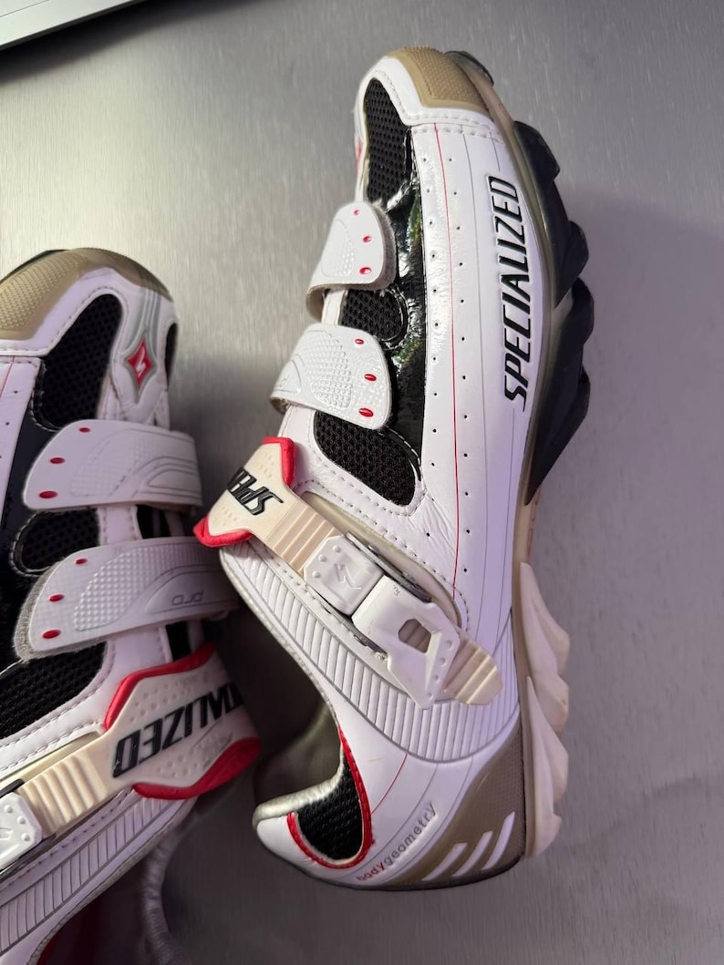 May include: White and black Specialized cycling shoes with three adjustable straps. The shoes have a red and beige accent colour scheme, with the word "SPECIALIZED" printed on the side.