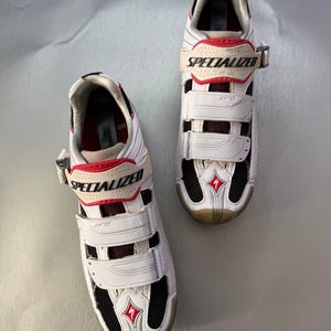 May include: Pair of white and red Specialized Pro cycling shoes. The shoes have three adjustable straps, black mesh vents, and the Specialized logo. The soles are a light tan colour. The shoes are designed for cycling.