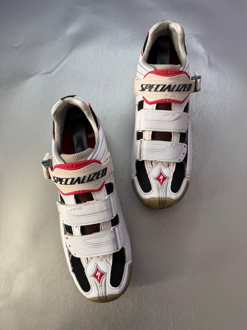 May include: Pair of white and red Specialized Pro cycling shoes. The shoes have three adjustable straps, black mesh vents, and the Specialized logo. The soles are a light tan colour. The shoes are designed for cycling.