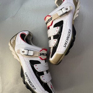 May include: Pair of white and black Specialized cycling shoes with red detailing. The shoes feature three Velcro straps and gold-coloured heels and toes. The word "SPECIALIZED" is printed on the side of each shoe.