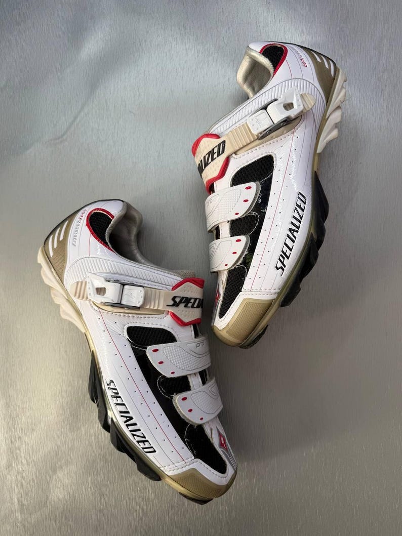 May include: Pair of white and black Specialized cycling shoes with red detailing. The shoes feature three Velcro straps and gold-coloured heels and toes. The word "SPECIALIZED" is printed on the side of each shoe.