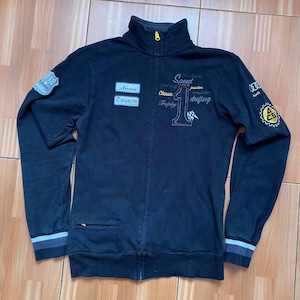 May include: Black zip-up jacket with racing-themed patches and embroidered details. Features the words "Speed", "Classic", and "Trophy". Includes a small yellow zipper and blue and white striped cuffs.