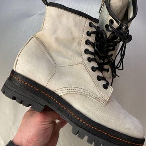 May include: Off-white suede ankle boot with black laces and a black sole. The boot features a zipper on the side and orange stitching around the sole. The boot is held by a person.