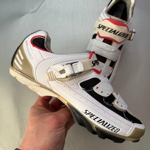 May include: White and black Specialized cycling shoes with red detailing. The shoes feature a gold heel and the word "SPECIALIZED" is printed on the side. They have adjustable straps and are designed for cycling.