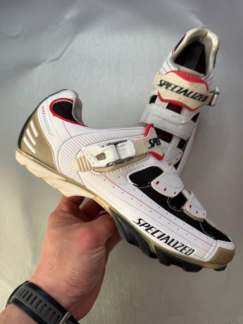 May include: White and black Specialized cycling shoes with red detailing. The shoes feature a gold heel and the word "SPECIALIZED" is printed on the side. They have adjustable straps and are designed for cycling.