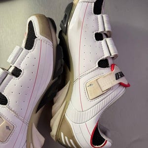 May include: Pair of white cycling shoes with beige accents and red detailing. The shoes have a hook-and-loop closure system and a black sole. The design includes mesh ventilation and a streamlined shape, suitable for athletic activities.