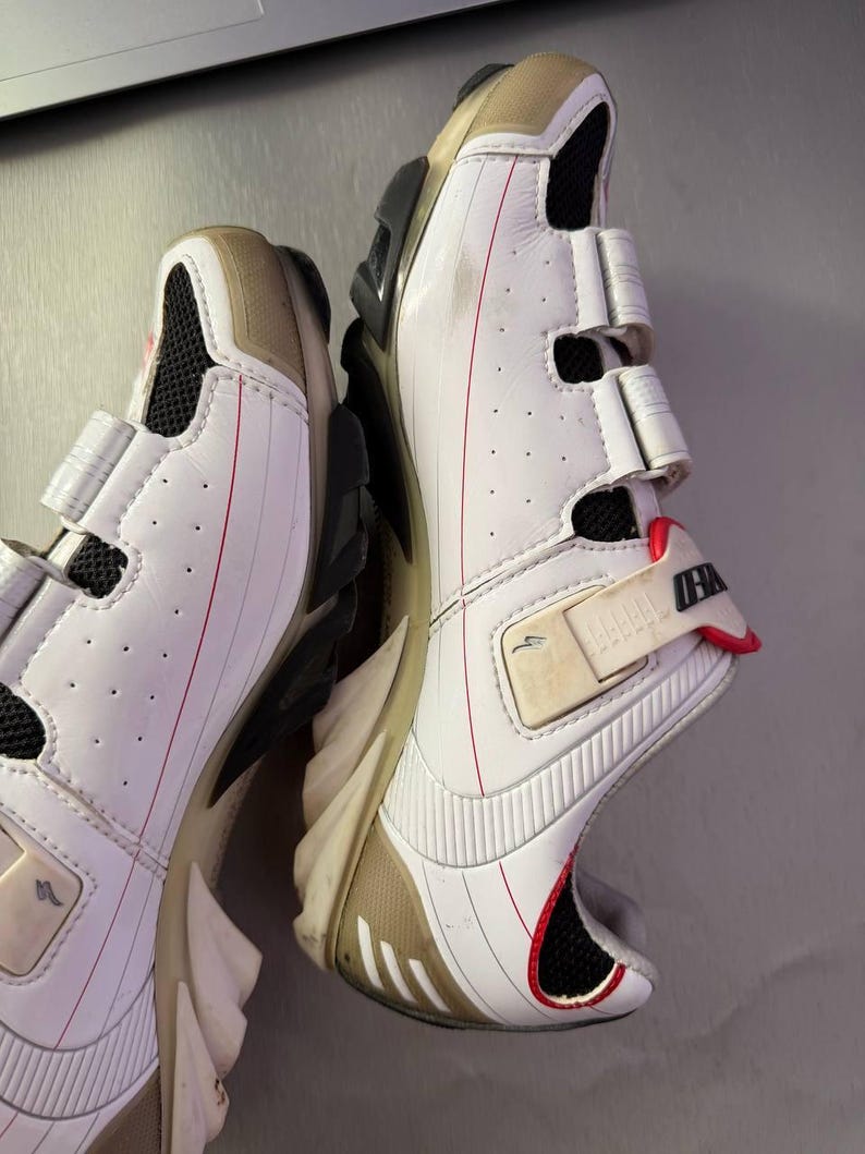 May include: Pair of white cycling shoes with beige accents and red detailing. The shoes have a hook-and-loop closure system and a black sole. The design includes mesh ventilation and a streamlined shape, suitable for athletic activities.