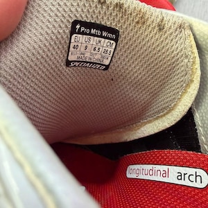 May include: Detail of a Specialized Pro Mtb Wmn shoe interior. The shoe features a red and grey design with a label showing size details: EU 40, US 9, UK 6.5, and 25.5 cm. The words "longitudinal arch" are printed on the shoe.