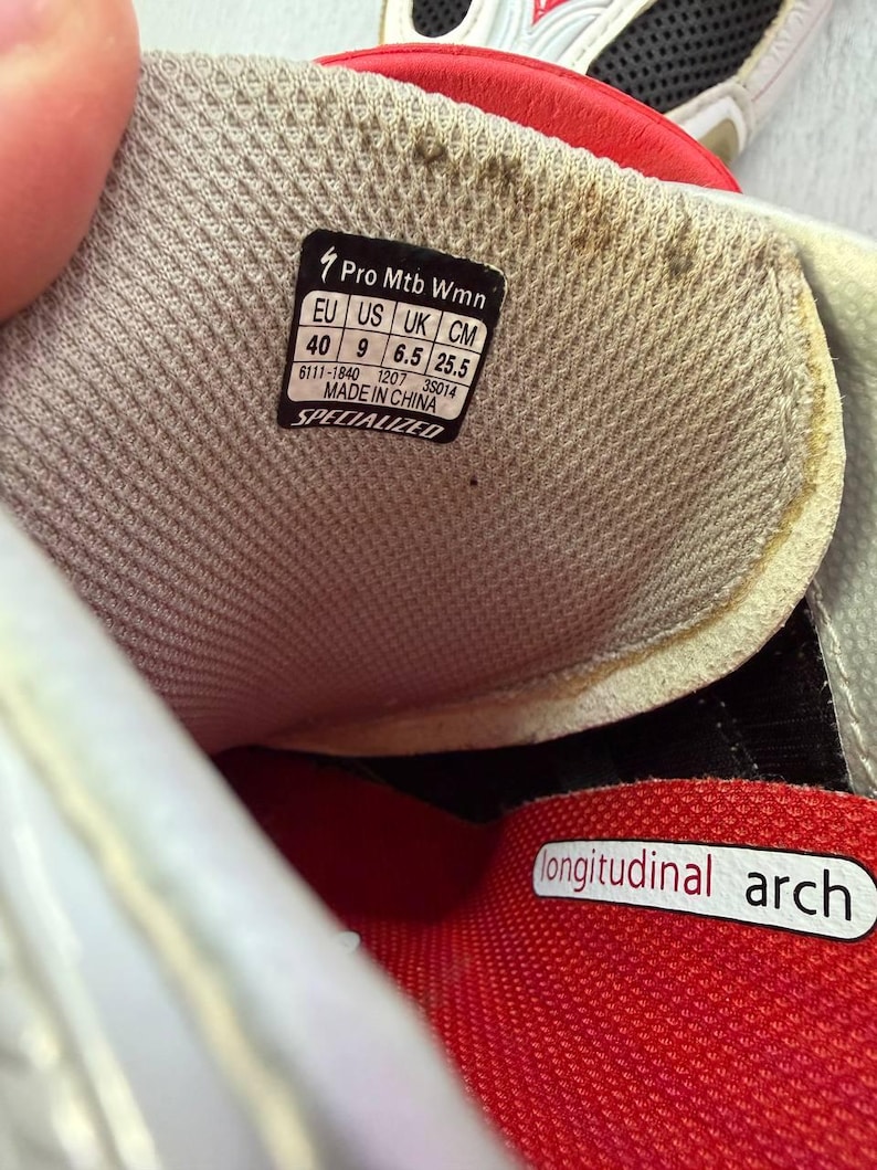 May include: Detail of a Specialized Pro Mtb Wmn shoe interior. The shoe features a red and grey design with a label showing size details: EU 40, US 9, UK 6.5, and 25.5 cm. The words "longitudinal arch" are printed on the shoe.