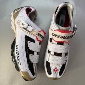 May include: Pair of white and black Specialized cycling shoes with red detailing. The shoes have adjustable straps, the word "SPECIALIZED" printed on the side, and a gold-coloured sole. Designed for cycling.