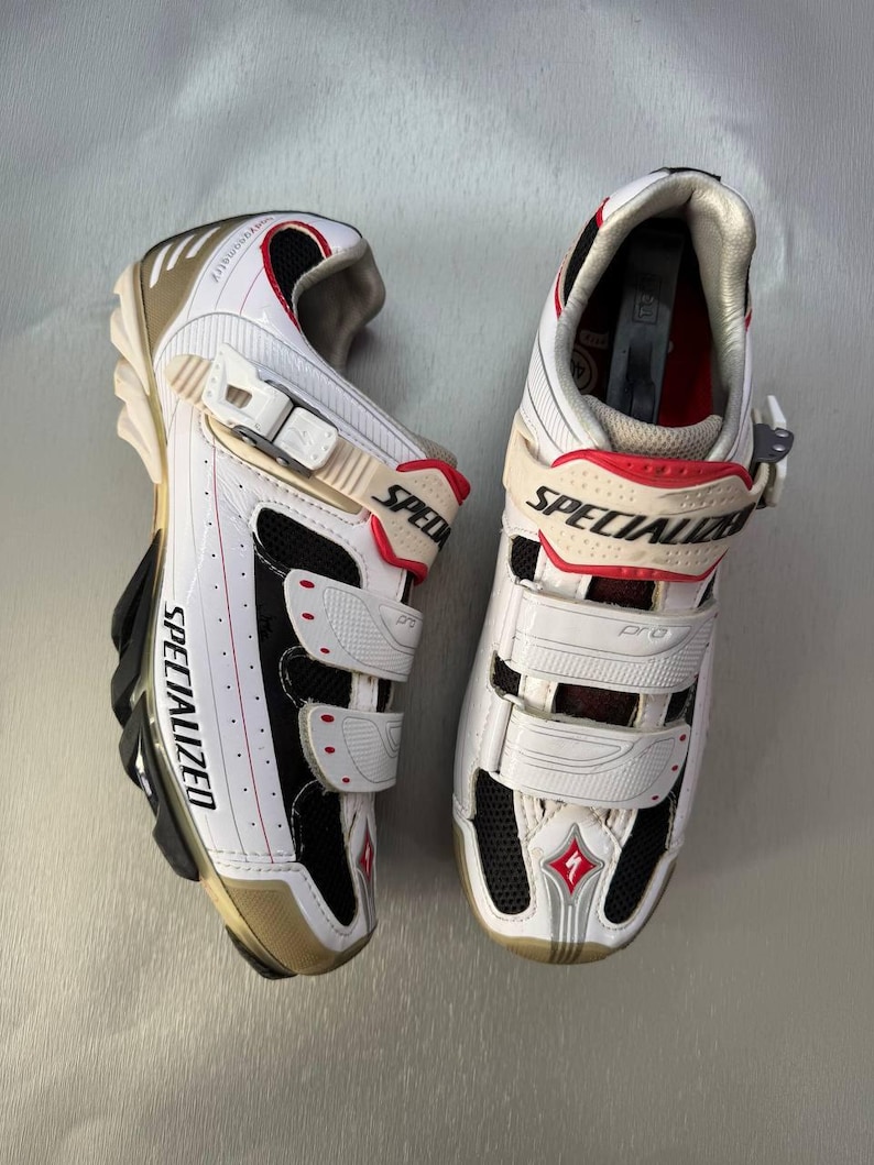 May include: Pair of white and black Specialized cycling shoes with red detailing. The shoes have adjustable straps, the word "SPECIALIZED" printed on the side, and a gold-coloured sole. Designed for cycling.