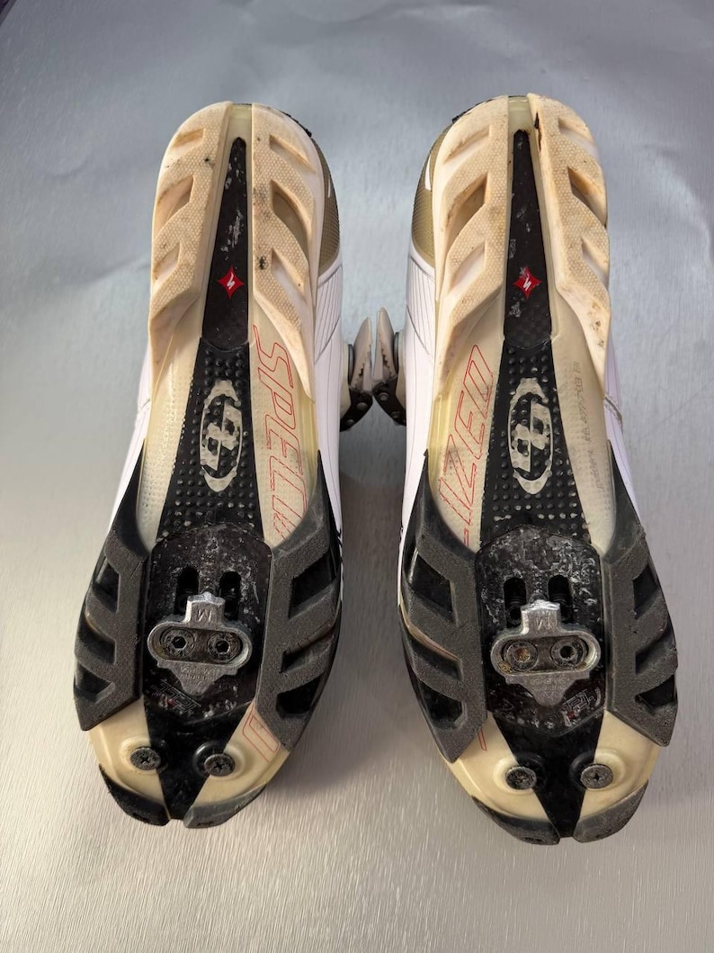 May include: Pair of white and black cycling shoes with a Specialized logo. The soles are visible, showing the cleat attachment points and the word "SPECIALIZED" in red. The shoes have a worn appearance.