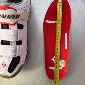May include: White and red Specialized cycling shoe next to a red insole being measured with a tape measure. The insole is approximately 25 cm long. The shoe has three white straps and the word "SPECIALIZED" is visible.