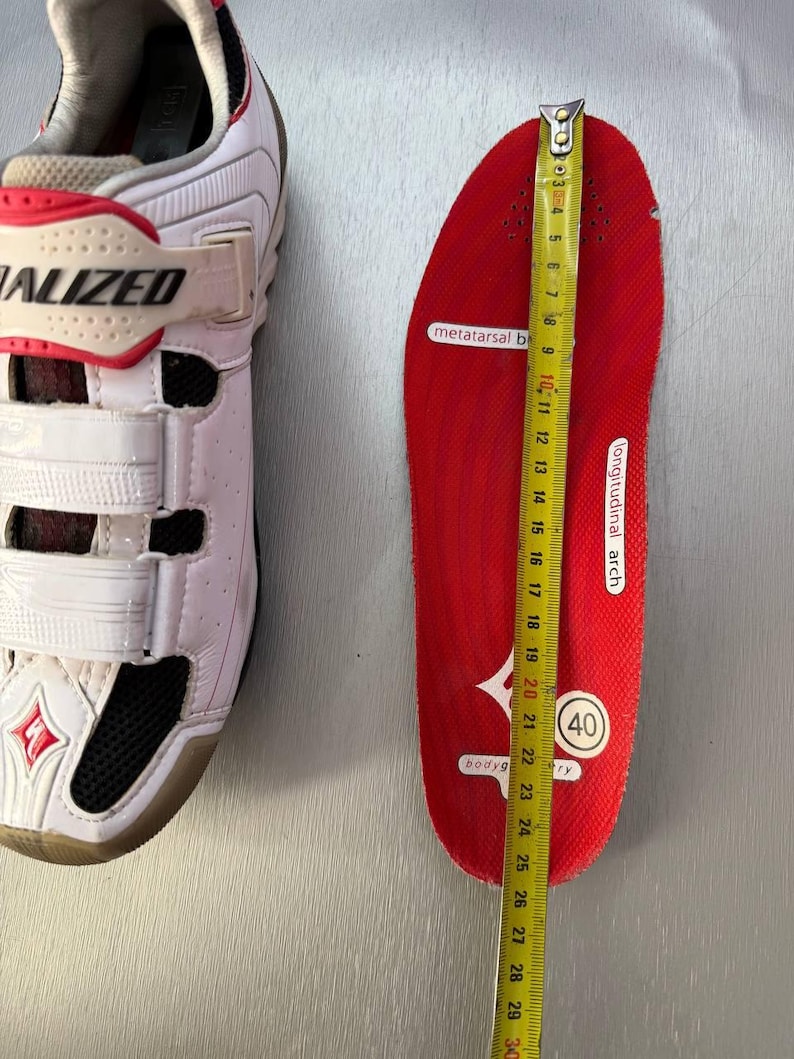 May include: White and red Specialized cycling shoe next to a red insole being measured with a tape measure. The insole is approximately 25 cm long. The shoe has three white straps and the word "SPECIALIZED" is visible.