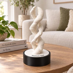 Könnte beinhalten: Eine weiße abstrakte Skulptur mit zwei ineinander verschlungenen, gerippten, spiralförmigen Formen, die von einer runden Basis aufsteigen. Die Basis ist weiß mit dem schwarzen Text Custom Text auf der Vorderseite und steht auf einem schwarzen Sockel.