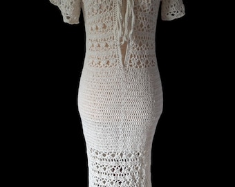 Könnte beinhalten: Cremefarbenes, handgefertigtes Häkelkleid, präsentiert auf einer Schaufensterpuppe. Das Kleid hat einen V-Ausschnitt mit Bindeband, kurze Ärmel und eine taillierte Silhouette, die am Saum ausgestellt ist. Das Lochmuster verleiht dem Kleid eine zarte Textur.