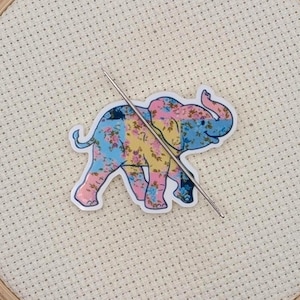 May include: A colorful elephant sticker with floral patterns in pink, yellow, and blue is centered on a cross-stitch hoop. A needle is inserted through the fabric, suggesting a craft project. The wooden hoop frames the off-white fabric.