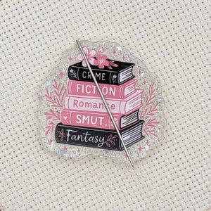 May include: A stack of books with the words "Crime", "Fiction", "Romance", "Smut", and "Fantasy" on the spines. The books are pink and black with a floral design. A needle is placed through the books, set in a wooden embroidery hoop.