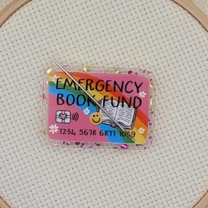 May include: A cross-stitch hoop with a needle piercing a clear acrylic card. The card features the text "EMERGENCY BOOK FUND" over a rainbow, a book, and a credit card design. The card also has the numbers 1234 5678 6877 1059.