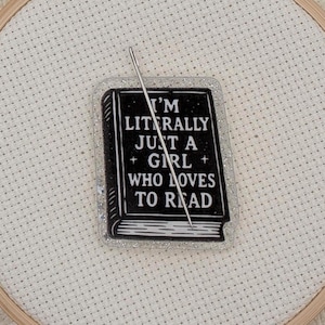 May include: A black and white book-shaped pin with the text "I'M LITERALLY JUST A GIRL WHO LOVES TO READ" is centered on a white fabric. A silver needle is inserted through the pin. The pin is surrounded by glitter and set in a wooden embroidery hoop.
