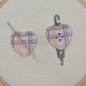 May include: A close-up of a cross-stitch project in a wooden hoop. Two heart-shaped buttons with a plaid pattern are visible, one with a needle through it. The buttons are peach, white, and lavender. The fabric is a light beige.