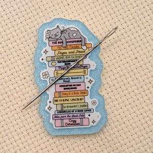 May include: A light blue enamel pin featuring a stack of books with a sleeping cat on top. The pin includes the text "THE BOOK WHISPERER" and other book titles. A silver needle is placed diagonally across the pin, set on a white fabric background.