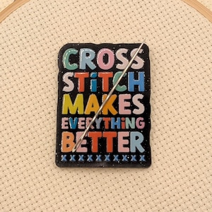 May include: A black pin with the text "CROSS STITCH MAKES EVERYTHING BETTER" in colorful letters. A needle is inserted through the pin, which is placed on a cross-stitch fabric stretched within a wooden embroidery hoop.