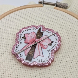 May include: A pink glittery cross-shaped embellishment with a pink bow and a needle. The cross is brown with a pink bow. The text on the embellishment reads "Clothed with strength and dignity. She laughs without fear of the future 31:25."