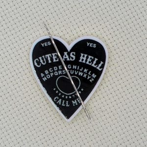May include: A cross-stitch project in a wooden embroidery hoop. The design features a black heart-shaped planchette with the words "CUTE AS HELL" and "CALL ME". A needle is inserted through the fabric.