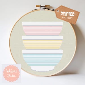 May include: A cross-stitch design featuring a stack of three mixing bowls in pastel colors: pink, yellow, and blue. The bowls have horizontal striped patterns. The design is displayed in a wooden embroidery hoop with a tag that says "PRINTED PATTERN".