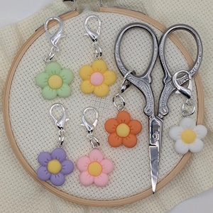 May include: A collection of colorful flower-shaped charms with silver clasps, alongside a pair of ornate embroidery scissors. The charms are in shades of green, yellow, orange, purple, pink, and white, each with a contrasting center.