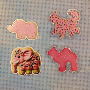 May include: Four animal-shaped needle minders. One is a pink elephant with sprinkles and a metal needle. Another is a pink elephant with small dots. A third is a pink cat with colorful dots and glitter. The fourth is a pink camel with glitter.