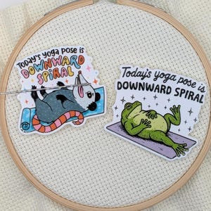 May include: Two stickers on a white fabric background. One sticker features a possum in a yoga pose with the text "Today's yoga pose is DOWNWARD SPIRAL". The other sticker shows a frog in a yoga pose with the same text.
