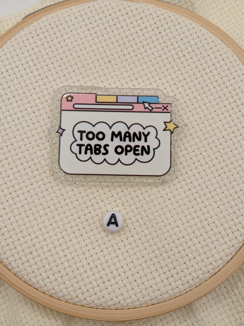 Peut inclure: &Eacute;pinglette transparente et scintillante en forme de fen&ecirc;tre de navigateur web avec le texte "TOO MANY TABS OPEN" dans une bulle. L'&eacute;pinglette a des accents roses, jaunes, bleus et violets pastel. Une petite perle blanche avec la lettre "A" est &eacute;galement visible.