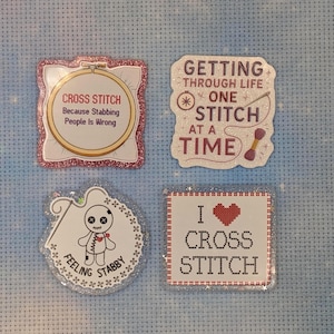 May include: Four glittery stickers with craft themes. One shows a hoop with text "CROSS STITCH Because Stabbing People Is Wrong". Another says "GETTING THROUGH LIFE ONE STITCH AT A TIME". A third features a voodoo doll with "FEELING STABBY". The last says "I [heart] CROSS STITCH".
