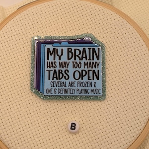 Peut inclure: Un cercle &agrave; broder avec un carr&eacute; bleu et scintillant portant le texte "MY BRAIN HAS WAY TOO MANY TABS OPEN. SEVERAL ARE FROZEN & ONE IS DEFINITELY PLAYING MUSIC." Une petite perle blanche avec la lettre "B" est &eacute;galement pr&eacute;sente.
