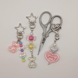 May include: A collection of keychains and a pair of silver scissors. The keychains feature star-shaped clasps, colorful beads, and charms including a teddy bear, candy-shaped beads, and a pink heart. The scissors have ornate handles.
